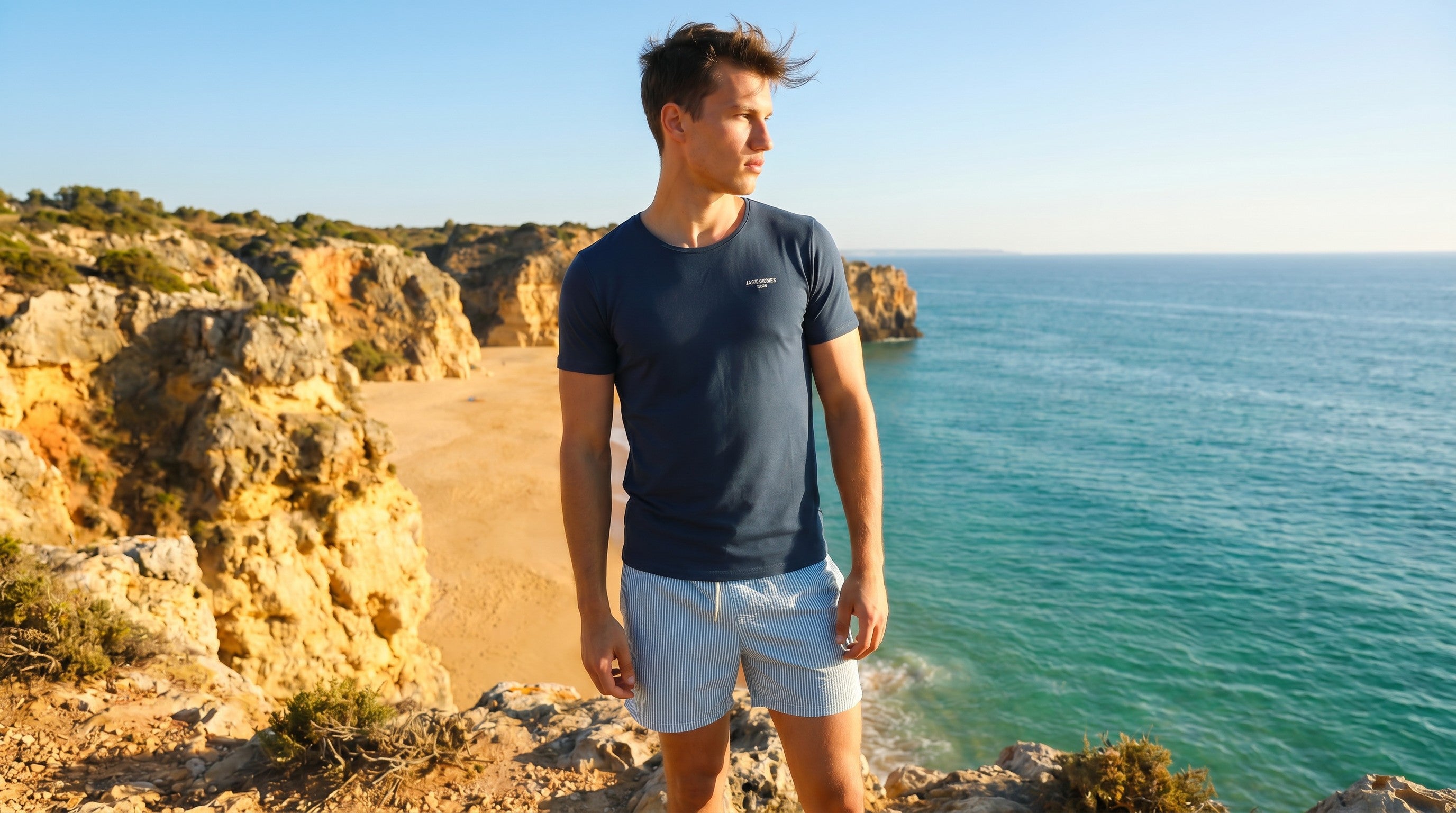 Man wearing a Jack Rabbit Swims navy swim tee standing on coastal cliffs with turquoise ocean views