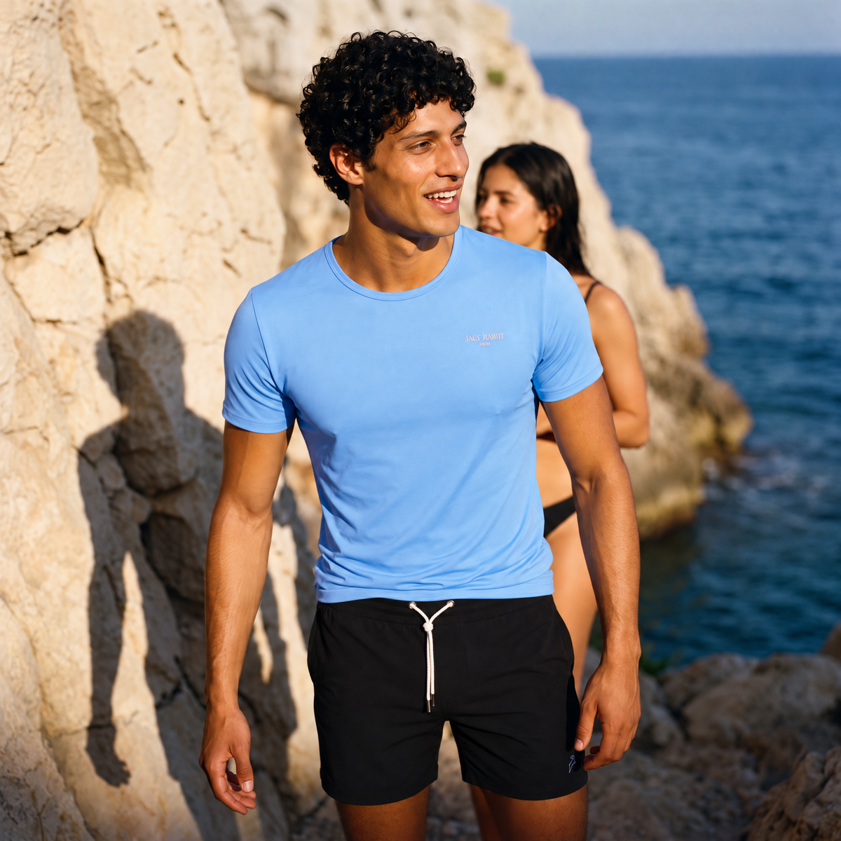 Jack Rabbit Swims Light Blue Swim Tee worn with black swim shorts on a rocky beach.