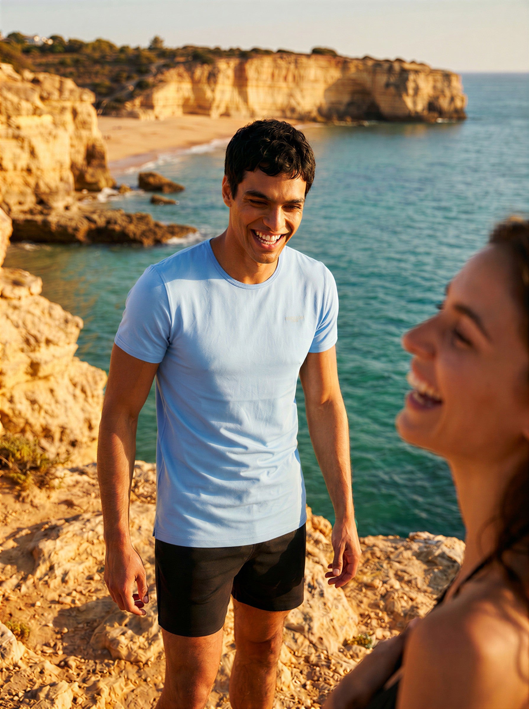 Man wearing a Jack Rabbit Swims light blue swim tee laughing with a woman on coastal rocks